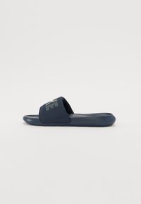 Sandale cu toc de culoare albastru marin, cu o talpă texturată și o curea largă care include un logo contrastant. Fabricate din material sintetic, concepute pentru confort.