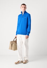 Blauer Strickpullover mit hohem Kragen, kombiniert mit weißen Hosen, braun-weißen Sneakers und einer beigen Tasche mit braunen Akzenten.