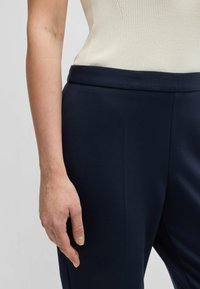 Pantaloni tailored blu navy con una texture liscia, caratterizzati da una vita pulita e una vestibilità aderente, abbinati a un top in crema a coste.