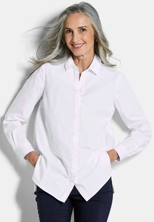Femme souriante aux longs cheveux gris portant une chemise blanche boutonnée et un pantalon sombre, debout les mains dans les poches devant un fond blanc.