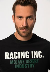 Camiseta negra de algodón con gran texto blanco y verde: "RACING INC. INDUSTRIA DEL DESIERTO MOJAVE". Diseño clásico de cuello redondo.