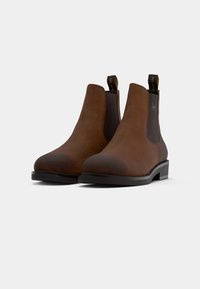 Botas Chelsea de cuero marrón con paneles laterales elásticos, puntas redondeadas y lengüetas. Suelas de goma negra resistentes, textura suave y detalles mínimos.