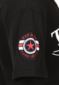 Musta puuvillainen t-paita lyhyillä hihoilla, jossa on pyöreä kirjailtu merkki. Merkillä on punainen tähti, "TOP GUN" ja "FLYING LEGEND" punaisella.