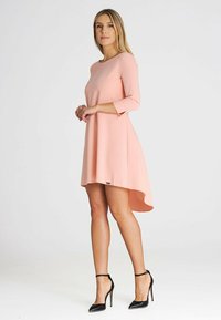 Rosa A-Linien-Kleid mit Dreiviertelärmeln, asymmetrischem Saum und glattem Stoff. Kombiniert mit schwarzen Stilettos.