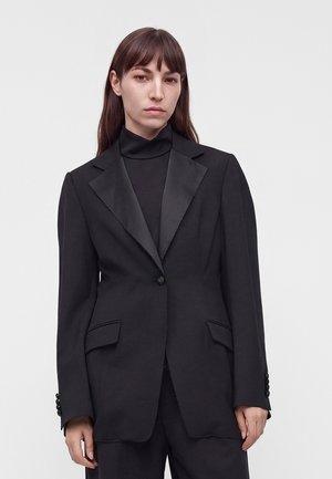 Donna con capelli castani lisci che indossa una giacca nera con revers in raso sopra un dolcevita nero, in piedi davanti a uno sfondo bianco uniforme.