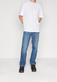 Hombre de pie con las manos detrás de la espalda, vistiendo una camiseta blanca sencilla, jeans azules, zapatillas negras y un collar delgado de plata.