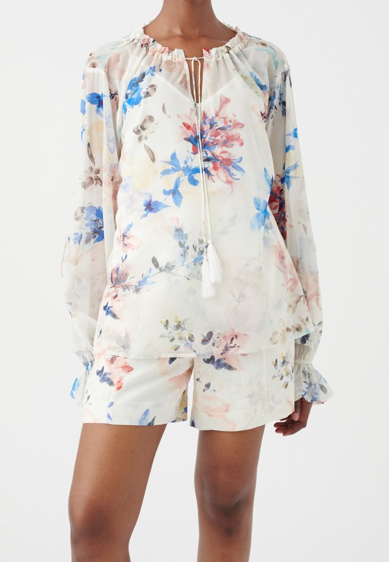 Femme portant un chemisier blanc transparent à manches longues avec des liens à pompons et un short fleuri assorti sur un fond uni.