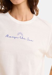 Biała bawełniana koszulka z niebieskim, kursywnym napisem "Zawsze Słońce" z prostą ilustracją słońca nad napisem. Okrągły dekolt, krótki rękaw.