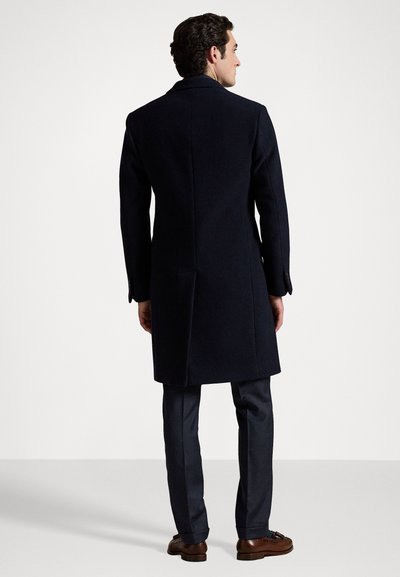 Palton din lână bleumarin cu un design ajustat, ventraș unic și guler cu dinți. Pur și simplu împreună cu pantaloni închiși la culoare și pantofi din piele maro.