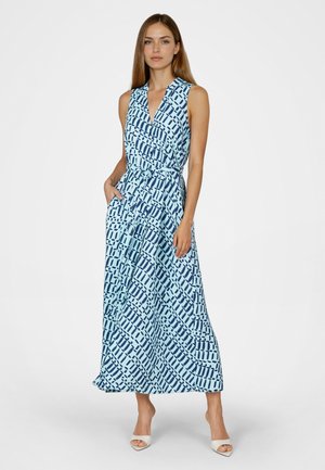 Woman wearing sleeveless blue maxi dress with navy geometric print, V-neck, tied waist, and white open-toe heels, standing with one hand in pocket.