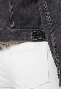 Černá džínová bunda s detailem knoflíků, kombinovaná s bílými kalhotami s kapsou na zadní straně. Tkanina má texturovaný vzhled.