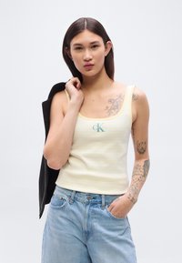 Κίτρινη και λευκή ριγέ μπλούζα χωρίς μανίκια με στρογγυλή λαιμόκοψη και κεντημένο λογότυπο. Συνδυασμένη με ανοιχτό μπλε τζιν παντελόνι. Απλός σχεδιασμός, απαλό ύφασμα.