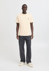 Cremefarvet t-shirt med korte ærmer og et lille grafik på brystet, parret med mørke denim straight-leg jeans og hvide sneakers.