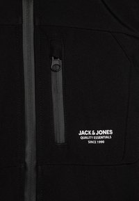 Veste noire avec une poche poitrine zippée, présentant des accents gris et un texte de marque blanc qui indique "JACK & JONES" avec "QUALITY ESSENTIALS."