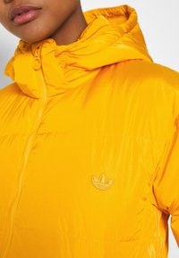 Gul puffjacka med dragkedja, elastisk huva och en liten broderad logotyp på bröstet. Skinande nylonmaterial med strukturerad yta.