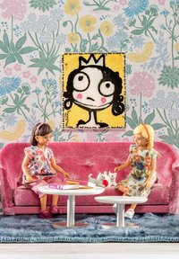 Two dolls sit on a pink, ribbed couch with floral-patterned dresses. A white table holds books and a red ball, with a plush rug beneath.