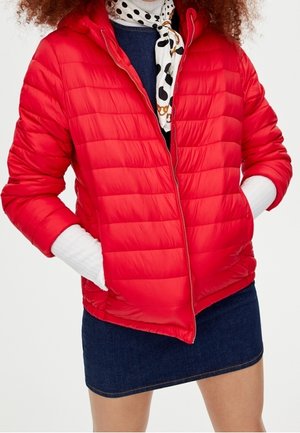 Woman wearing bright red quilted jacket, white ribbed sweater sleeves visible, dark denim skirt, and patterned neck scarf with polka dots and chains.