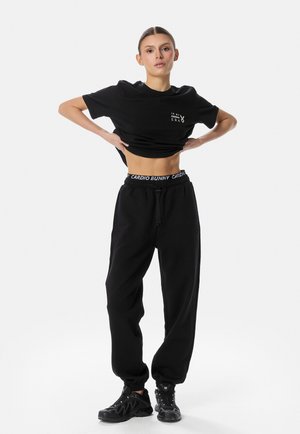 Kvinde i sorte træningsbukser og t-shirt løfter t-shirten for at afsløre maven, iført sorte sneakers, stående mod en ensfarvet hvid baggrund.