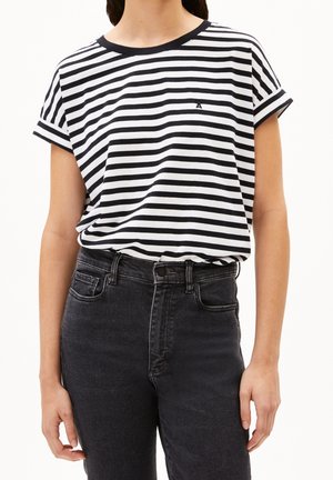 Femme portant un t-shirt à rayures horizontales noires et blanches à manches courtes, rentré dans un jean noir taille haute, debout avec les bras détendus.
