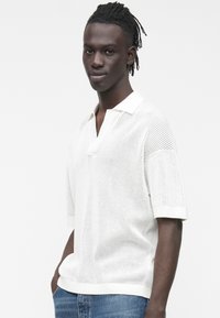 Biała dzianinowa koszulka polo z krótkimi rękawami, z klasycznym kołnierzykiem i fakturowanym wzorem, zestawiona z niebieskimi dżinsami.