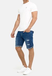 Hombre con una camiseta blanca, pantalones cortos de mezclilla desgastados y arremangados de color azul, y zapatillas blancas, de pie con una mano en el bolsillo.