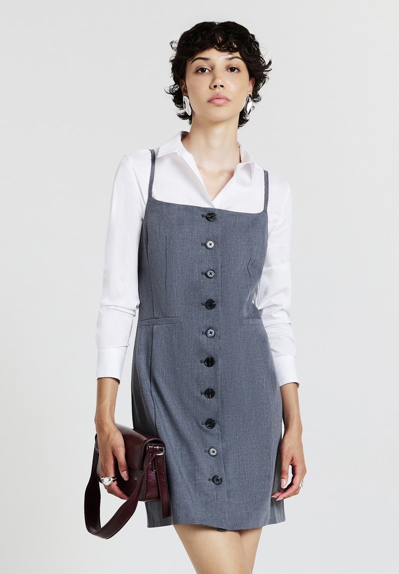 Abito grigio con scollo a camicia e spalline sottili, caratterizzato da bottoni neri sul davanti, abbinato a una camicia bianca e una borsa bordeaux.