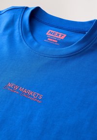 Blå bomulds-t-shirt med afslappet pasform, der har "NEW MARKETS" i pink skrifttype. Mærket viser mærket "NEXT," størrelse M, og "Fremstillet i Bangladesh."