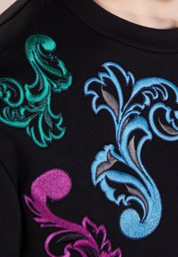 Tissu noir présentant une broderie complexe aux motifs tourbillonnants en vert, bleu et violet, mettant en valeur des textures variées et des couleurs vives.