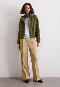 Oliwkowa kurtka z kapturem z dużymi guzikami i przednimi kieszeniami, nałożona na jasnoniebieski sweatshirt, w zestawie z beżowymi szerokimi spodniami.