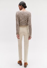 Beige op maat gemaakte broeken met achterzakken, gecombineerd met een gestructureerde bruine gebreide trui. Het model draagt bruine schoenen met zwarte accenten.