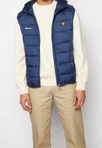 Tmavě modrá prošívaná vesta s kapucí, se zapínáním na přední zip a vyšívaným logem na levé straně hrudi. Nositelná přes krémově zbarvenou mikinu.