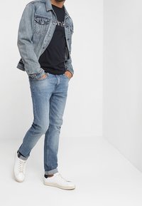 Mann trägt eine hellblaue Jeansjacke, ein schwarzes T-Shirt, hellblaue Jeans mit Aufschlägen und weiße Sneakers, steht vor einer weißen Wand und einem weißen Boden.