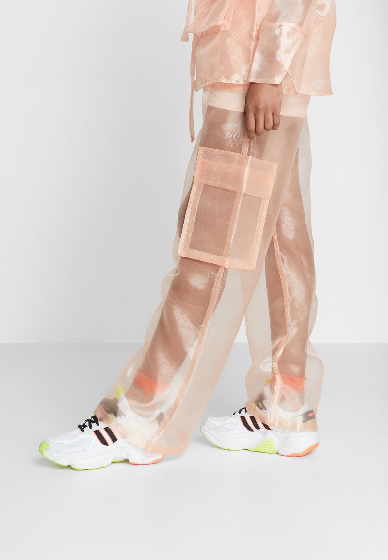 Person med genomskinliga persikofärgade byxor med stora sidofickor ovanför en liknande underlayer och vita sneakers med neonkonturer.