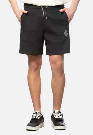 Mann trägt schwarze Shorts mit Kordelzug und Logo am linken Bein sowie schwarze Sneakers mit weißen Sohlen und Schnürsenkeln, Hände in den Taschen.