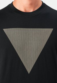 Camiseta negra con un gráfico de triángulo invertido a rayas beige que contiene el texto tenue "GUESS" en el pecho.
