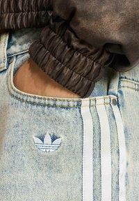 Polsino di giacca in pelle marrone con bordo arricciato accanto a shorts in denim azzurro chiaro con logo Adidas bianco e tre strisce bianche sul lato.