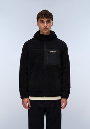 Jeune homme portant une veste polaire noire à fermeture éclair avec capuche et patch logo, debout devant un fond clair uni.