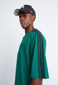 Grøn kortærmet T-shirt med sorte striber på skulderne og logo-detalje. Båret med en sort tætsiddende kasket, der viser en afslappet stil.