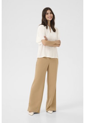 Blusa bianca a maniche corte con righe verticali; pantaloni larghi color beige chiaro; il modello posa con le braccia incrociate, indossando scarpe color crema.