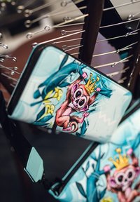 Péněženka na zip s ilustrací opice s korunou, umístěná na modro-zeleném abstraktním pozadí. Hladký povrch, živé barvy.