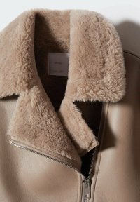 Veste en cuir beige avec un grand col en shearling doux et moelleux, et une fermeture éclair métallique partiellement ouverte à l’avant.
