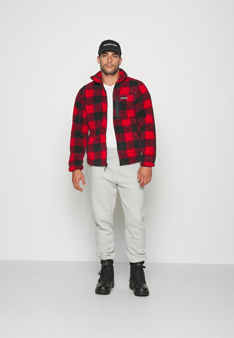 Veste en polaire à carreaux rouges et noirs, pantalon de survêtement gris et bottes noires. La veste dispose d'une poche zippée et d'un tissu texturé.