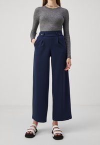 Navy wide-leg trousers with pleats, high waist, two buttons, and side pockets. Paired with a gray ribbed long-sleeve top and black sandals.