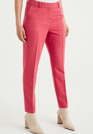 Femme portant un pantalon slim rose corail avec plis devant, poches latérales et bottines beiges à talons, debout sur un fond uni.