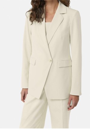 Kobieta ubrana w kremową, dopasowaną marynarkę i spodnie z wysokim stanem oraz top z kwadratowym dekoltem, stojąca na jednolitym tle.
