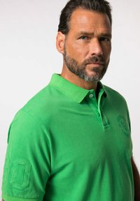 Camiseta polo verde de algodón con cuello, que cuenta con un logo bordado y el número "80" en la manga izquierda. Textura suave.