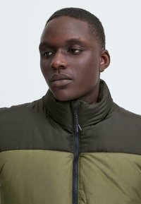 Olivegrüne gepolsterte Jacke mit hohem Kragen und Reißverschluss. Das zweifarbige Design hat einen dunkleren oberen Bereich und einen helleren unteren Bereich.
