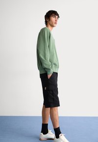 Green fleece sweatshirt, black cargo shorts, white sneakers. Features relaxed fit, ribbed cuffs, and a logo patch on shorts.