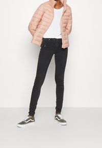 Helle rosa gepolsterte Jacke mit hohem Kragen, kombiniert mit einem weißen T-Shirt, schwarzen engen Jeans und olivgrünen Turnschuhen mit weißen Akzenten.