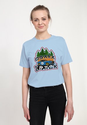 T-shirt bleu clair à manches courtes présentant un design graphique avec des arbres verts et une voiture bleue, accentué par des éléments de texte noirs et rouges.